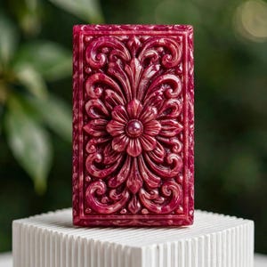 May include: A rectangular bar of deep magenta soap featuring an intricate, raised relief pattern of symmetrical floral and scrolling acanthus leaf motifs. The surface has a glossy, marbled finish with lighter pink highlights throughout the design.