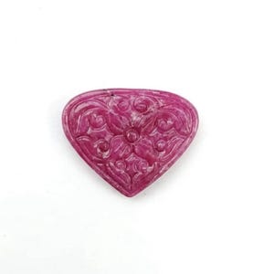 Puede incluir: Una piedra preciosa en forma de corazón de color magenta intenso con un intrincado patrón floral tallado en su superficie. La piedra tiene un acabado liso y pulido con detalles en relieve que definen los diseños simétricos de pétalos en espiral.