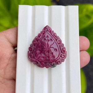 May include: A teardrop-shaped deep magenta gemstone pendant featuring an intricate carved floral pattern on its surface. The stone has a decorative scalloped border with ornate scrollwork details, showcasing a polished finish and rich colour.