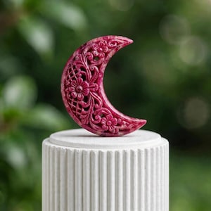 Puede incluir: Una talla en forma de media luna de un tono magenta intenso, con intrincados detalles en relieve de flores y patrones de celosía. El objeto se exhibe sobre un pedestal cilíndrico blanco con estrías verticales, mostrando su detallada artesanía.