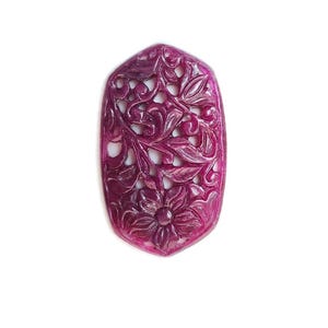 Könnte beinhalten: Ein tief magentafarbener, ovaler Edelsteinanhänger mit einem filigranen, handgeschnitzten Durchbruchdesign aus Blumenmotiven und geschwungenen Rankenmustern. Die Oberfläche weist ein poliertes, durchscheinendes Finish in dunklen Rosatönen auf.