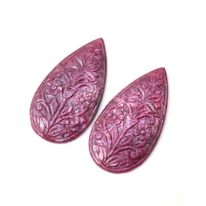 Puede incluir: Dos piedras preciosas de rubí en forma de lágrima con intrincados patrones florales tallados a mano. Cada piedra de color rojo magenta intenso muestra una superficie lisa y pulida con grabados detallados de flores y tallos en la cara frontal.