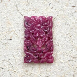 May include: A rectangular pendant carved from deep magenta ruby gemstone featuring an intricate openwork floral design with a prominent central flower and symmetrical leaf motifs throughout the surface, showcasing natural dark inclusions.