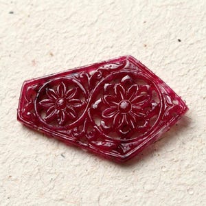 Puede incluir: Una talla de piedra preciosa de color rojo intenso, translúcida y con forma de cometa, que presenta un intrincado diseño calado de dos motivos florales estilizados conectados por enredaderas. La superficie está pulida y muestra un tono carmesí.