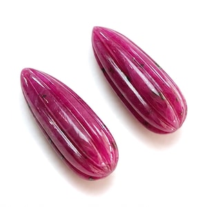 May include: Two teardrop-shaped ruby gemstones with a deep magenta hue and a polished, vertically fluted surface. Each stone features natural dark inclusions and a smooth, glossy finish, showcasing a distinct elongated form with rounded ends.