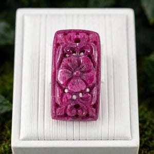 May include: A rectangular, deep magenta-colored gemstone pendant featuring an intricate, hand-carved floral motif with openwork detailing. The surface of the stone displays a natural, crystalline texture with varied shades of pink and red throughout.