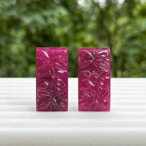 May include: Two rectangular, deep pink gemstone pieces featuring intricate, hand-carved floral patterns across their surfaces. Each piece displays a polished finish with visible carved petals and leaves, showcasing a consistent vibrant magenta hue throughout.