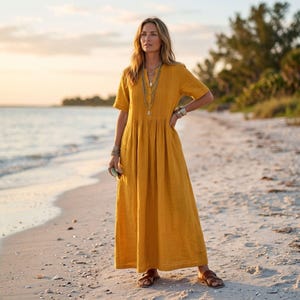 May include: A mustard yellow maxi dress featuring short sleeves, a V-neckline, and vertical pleats along the bodice. The dress has a loose, flowing skirt that reaches the ankles, paired with layered silver necklaces and brown leather sandals.