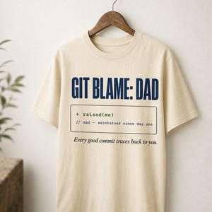 Puede incluir: Una camiseta beige de manga corta con texto azul oscuro que dice GIT BLAME: DAD. Debajo, un recuadro rectangular contiene texto estilo código: + raised(me) // dad - maintainer since day one. El texto inferior dice: Every good commit traces back to you.