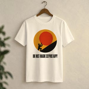 以下が含まれることがあります： クリーム色の半袖Tシャツ。黄色い太陽を背景に、黒い丘の上へ大きな赤い円を押す黒い人物のグラフィックが描かれています。グラフィックの下には「ONE MUST IMAGINE SISYPHUS HAPPY」というテキストがプリントされています。