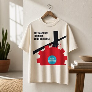 以下が含まれることがあります： 「THE MACHINE FINISHES YOUR SENTENCE」のテキストが入ったクリーム色のTシャツ。グラフィックは、"AUTO COMPLETE"と書かれた青い円が付いた赤い歯車が特徴です。黒い手が歯車の上に描かれています。
