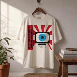 May include: Off-white t-shirt with a graphic of a TV screen with a blue eye, red rays, and the text "WHO REVIEWS/ THE REVIEWER" and "YOU WATCH IT/ IT WATCHES YOU". The shirt is displayed on a wooden hanger.