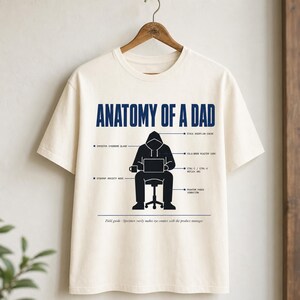 May include: An off-white short-sleeve t-shirt featuring a dark blue and black graphic titled Anatomy of a Dad. The design shows a hooded figure sitting at a desk with a laptop, surrounded by labels like Imposter Syndrome Gland and Cold-Brew Reactor Core.