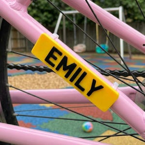 Peut inclure: Une plaque signalétique rectangulaire jaune avec le nom EMILY imprimé en lettres capitales noires grasses. La plaque est fixée sur le cadre métallique rose clair d'un vélo à l'aide de deux attaches en plastique transparent, positionnée horizontalement.