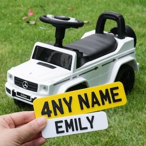 May include: A white toy SUV with a black steering wheel and seat, featuring a Mercedes-Benz logo on the front grille. In the foreground, two rectangular number plates are held, one yellow with the text 4NY NAME and one white with EMILY.