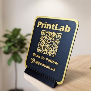 May include: A rectangular black sign with a gold border and gold raised lettering. The sign features the text PrintLab, a QR code, the text Scan to Follow, an Instagram icon, and the handle @printlab.uk. It rests on a black stand.