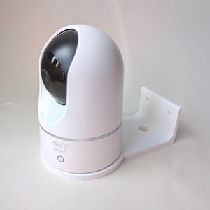 Könnte beinhalten: Eine weiße Eufy-Sicherheitskamera mit schwarzem Objektiv und Montagehalterung. Die Kamera hat eine abgerundete Oberseite und einen kreisförmigen Sockel. Der Markenname und ein Power-Button sind sichtbar.