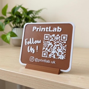May include: A rectangular brown sign with a white border and stand, featuring the white text PrintLab, Follow Us!, and @printlab.uk, along with a white QR code. The sign is designed for display on a flat surface to provide social media information.