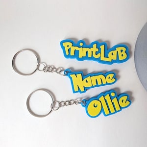 May include: Three custom keychains featuring yellow lettering on a blue background. The top keychain reads PrintLab, the middle reads Name, and the bottom reads Ollie. Each keychain includes a silver metal split ring attached by a short chain.