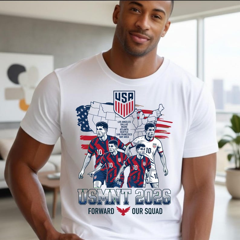 以下が含まれることがあります： アメリカの地図、サッカーアメリカ代表のエンブレム、背番号10のユニフォームを着た4人のサッカー選手が描かれた白い半袖Tシャツ。テキストには「USMNT 2026」、「Forward Our Squad」、開催都市のリストが含まれています。