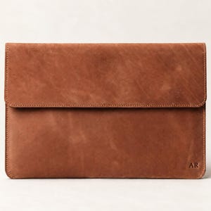 Puede incluir: Una funda rectangular de cuero marrón para portátil con cierre de solapa. La superficie tiene una textura mate y natural con variaciones de grano visibles. La esquina inferior derecha presenta las letras mayúsculas AR en relieve.