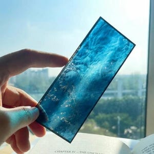 May include: A rectangular bookmark featuring a translucent blue design with swirling patterns and light-catching textures. The bookmark has a thin black border and displays small white text along the side, held against a bright window.