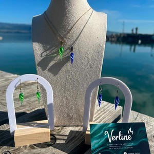 Può includere: Esposizione di gioielli in vetro, tra cui collane con ciondoli e orecchini. Le collane hanno ciondoli verdi e blu. Gli orecchini sono esposti su archi bianchi. È visibile anche un biglietto da visita con il nome "Verline".
