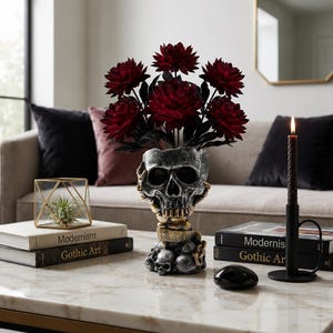 Pode incluir: Um vaso decorativo em forma de caveira com acabamento cinza e dourado envelhecido, contendo um buquê de flores vermelho-escuras. O vaso repousa sobre uma base de caveiras menores entre pilhas de livros e uma vela preta.