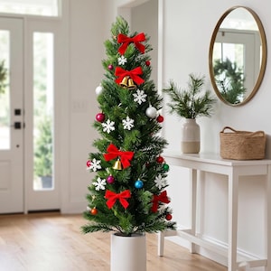 May include: A tall, slender artificial Christmas tree in a white cylindrical pot, decorated with red velvet bows, gold bells, white snowflake ornaments, and various colored baubles including red, silver, green, and blue spheres.