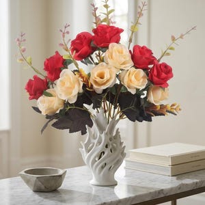 May include: A white ceramic vase with a textured, swirling, flame-like design holds an arrangement of artificial flowers. The bouquet consists of deep red and cream-colored roses mixed with dark green leaves and thin, light-colored sprigs.