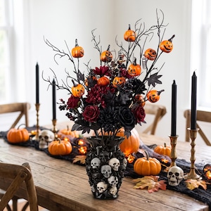 May include: A black vase decorated with multiple white and grey skull figures, filled with an arrangement of dark red and black artificial flowers, bare black branches, and small orange jack-o'-lantern ornaments with carved faces.