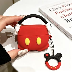 May include: A red silicone protective case for wireless earbuds featuring a black handle with white glove accents, two yellow oval buttons on the front, and a matching black and red circular ring charm with a small red heart detail attached.