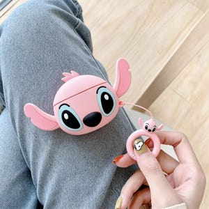 May include: A light pink silicone protective case for wireless earbuds, shaped like the character Angel with large ears, black eyes, and a black nose. A matching pink ring charm with a small Angel figure is attached by a thin cord to the main case.