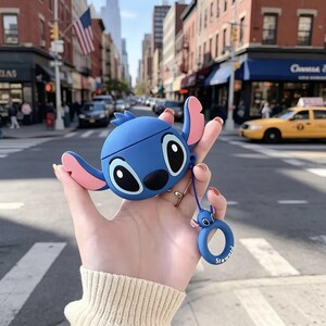 May include: A blue silicone protective case for wireless earbuds shaped like the character Stitch, featuring large black eyes, a black nose, and pink inner ears. A matching blue circular ring charm with a small Stitch head is attached by a cord.
