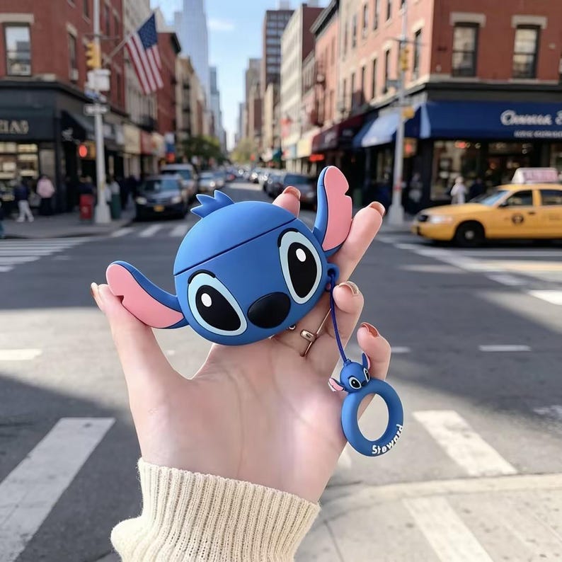 May include: A blue silicone protective case for wireless earbuds shaped like the character Stitch, featuring large black eyes, a black nose, and pink inner ears. A matching blue circular ring charm with a small Stitch head is attached by a cord.