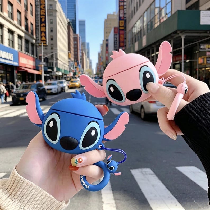 May include: Two silicone protective cases for wireless earbuds, shaped like cartoon characters. One case is blue with pink inner ears and large black eyes, the other is pink with similar features. Each includes a matching character ring charm.