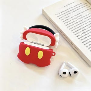 May include: A red silicone protective case for wireless earbuds, designed with two yellow oval accents on the front and white glove-like details on the sides. The case is open, revealing a white interior, and is accompanied by two white earbuds.