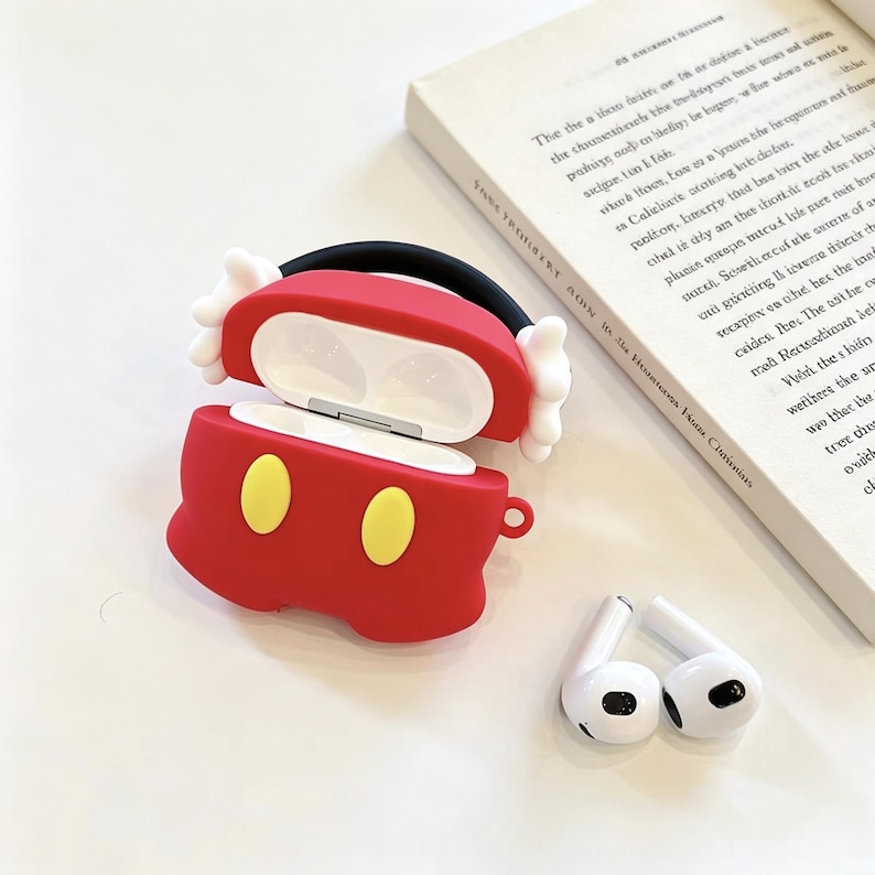 May include: A red silicone protective case for wireless earbuds, designed with two yellow oval accents on the front and white glove-like details on the sides. The case is open, revealing a white interior, and is accompanied by two white earbuds.