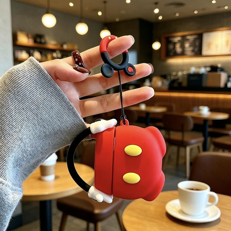 May include: A red silicone protective case for wireless earbuds featuring a cartoon-inspired design with two yellow oval accents, white glove-like details, and a black strap. The case includes a matching red and black circular ring attachment.
