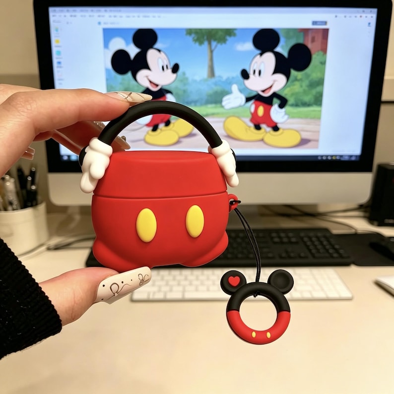 May include: A red silicone protective case for wireless earbuds, designed to resemble Mickey Mouse shorts with two yellow oval buttons. The case features a black handle with white gloved hands at the base and a matching black and red ring charm.