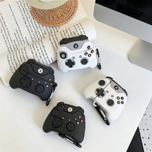 Può includere: Quattro custodie protettive in silicone per auricolari wireless, progettate per assomigliare a controller di gioco. Due custodie sono nero opaco e due sono bianche con accenti grigi. Ogni custodia presenta un D-pad, levette analogiche e pulsanti A, B, X, Y, con un moschettone in metallo.
