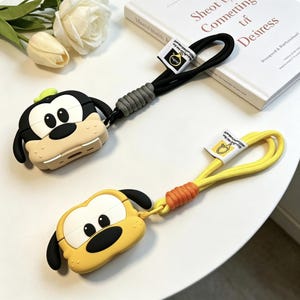Peut inclure: Deux étuis en silicone pour écouteurs en forme de têtes de chien de dessin animé. Un étui est noir et beige avec un chapeau vert et une dragonne noire. L'autre est jaune et noir avec une dragonne jaune. Les deux ont de petites étiquettes.