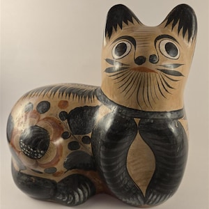 May include: A ceramic cat figurine featuring a tan base color with hand-painted black and brown floral and leaf motifs. The cat has large, wide-set eyes, pointed ears, and detailed black line work defining its face, whiskers, and body patterns.