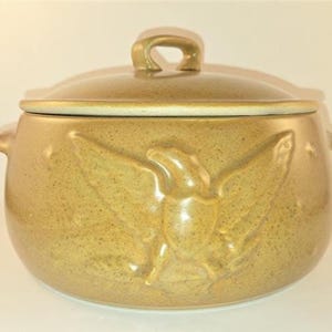 Pode incluir: Uma caçarola redonda de cerâmica em tom castanho-claro salpicado com tampa a condizer. O exterior apresenta um design de águia em relevo na parte frontal. O recipiente inclui duas pequenas pegas laterais e uma pega central na tampa.