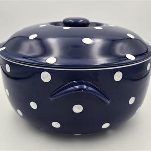 May include: A deep navy blue ceramic serving bowl with a matching lid, featuring a repeating pattern of white polka dots. The bowl has two integrated side handles and a rounded knob on the center of the lid, with a smooth, glossy finish.
