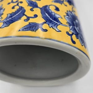 May include: A cylindrical ceramic vessel featuring a vibrant yellow background decorated with a repeating pattern of deep blue floral motifs and leafy vines. The base of the object is white, and the interior is a smooth, neutral gray finish.