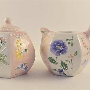Könnte beinhalten: Eine Zuckerdose aus Keramik mit Deckel und ein passendes Milchkännchen. Beide Teile haben eine sechseckige Form, eine zartrosa und weiße Farbpalette sowie handgemalte Blumenmotive in Violett, Gelb, Rosa und Grün mit erhabenen Perlendetails.