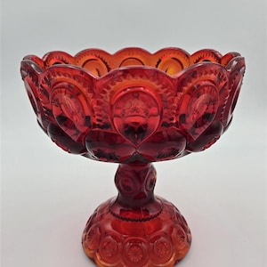 May include: A ruby red glass compote bowl featuring a repeating moon and star pattern. The bowl has a scalloped rim and sits on a pedestal base. The pressed glass design includes circular indentations and starburst motifs across the exterior surface.