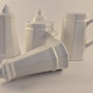 Könnte beinhalten: Ein Set aus vier weißen Keramik-Küchenartikeln, bestehend aus zwei hohen, konischen Salz- und Pfefferstreuern, einer kleinen Zuckerdose mit Deckel und seitlichen Griffen sowie einem Milchkännchen mit gebogenem Henkel, alle mit glatter Oberfläche.