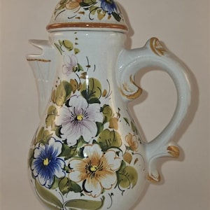 May include: A ceramic pitcher with a lid, featuring a white glazed finish and hand-painted floral motifs in shades of blue, yellow, and green. The vessel has a curved handle, a flared base, and a spout, with visible crackle glaze texture.